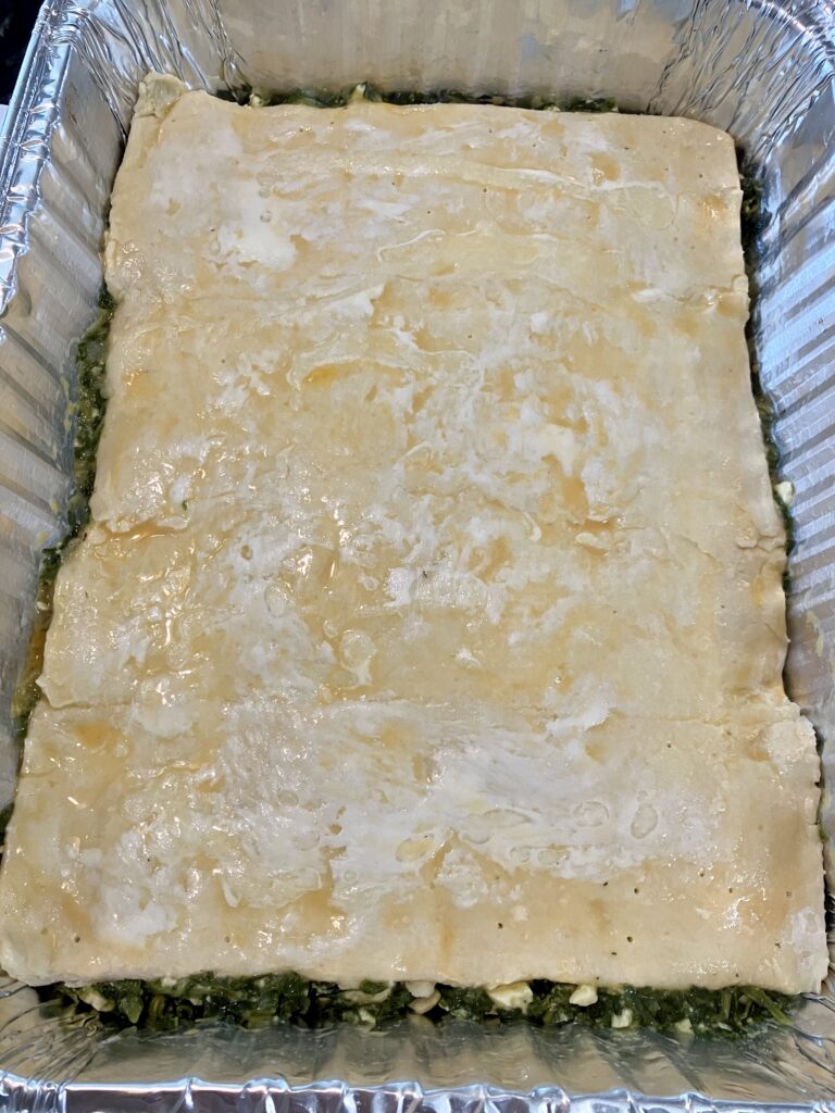 Top the spanakopita filling with the top layer of puff pastry. 